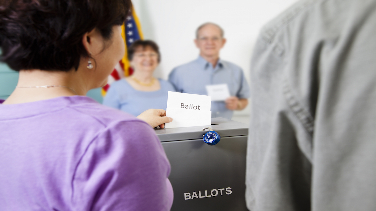 Multi-ethnic, mixed age group of people voting in the November USA elections at their local polling station. The voters stand in line at the local polling station to cast their ballot. Ballot boxes in a row.