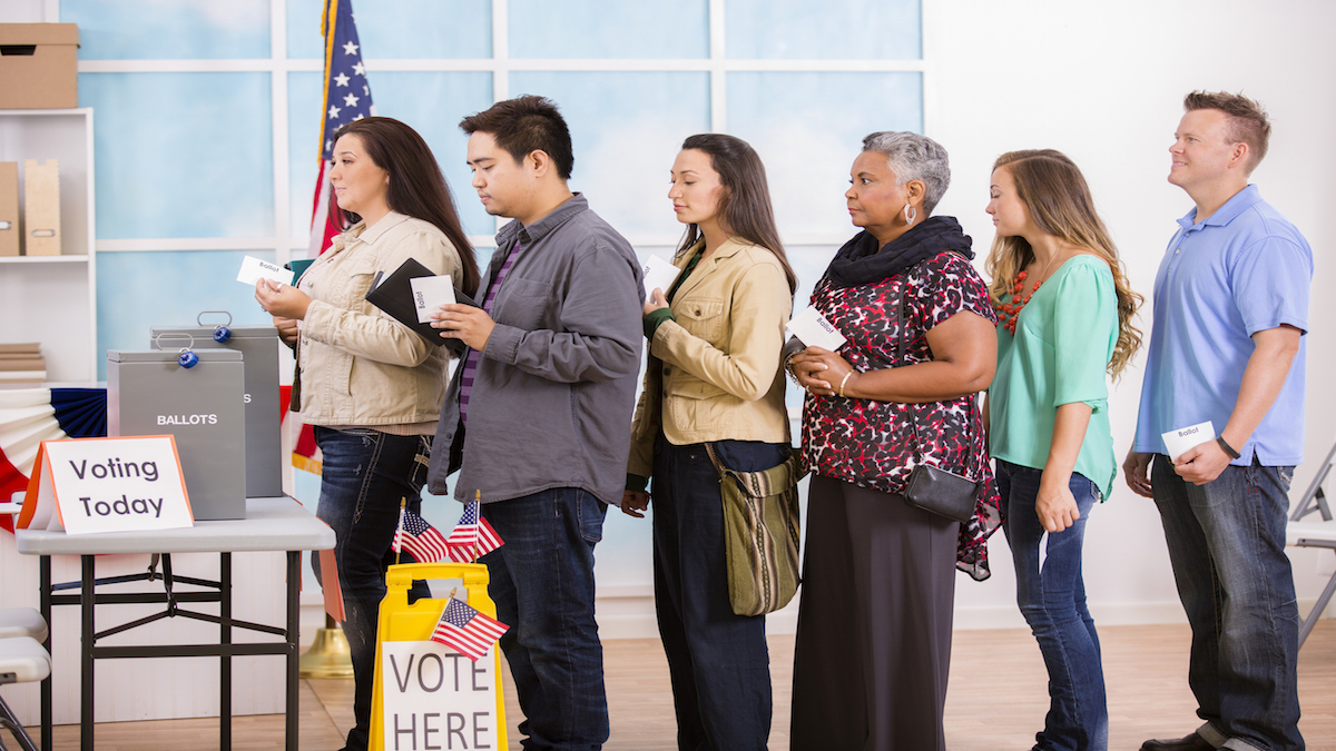 Multi-ethnic, mixed age group of USA citizens stand in line to cast their ballot in the November US elections at a local polling station. A table with ballot boxes, and "Voting Today" signs to left. USA flag in background.